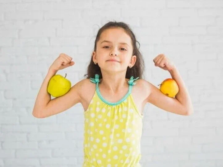 Menina sorridente fazendo pose de força com os braços flexionados, segurando uma maçã em cada braço, em frente a uma parede branca.